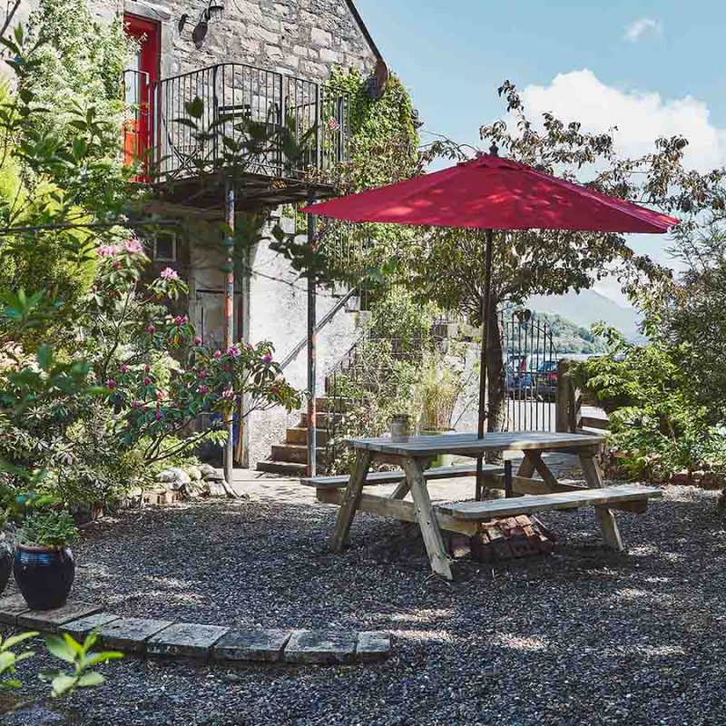 A beautiful lush courtyard with a picnic table with a red umbrella and a balcony off the apartment overlooking the courtyard at Lochview Apartment part of Brambles of Inveraray Hotel in Scotland
