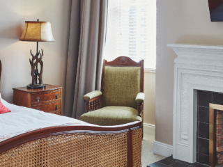 An antique rattan double bed, an antique armchair by a fireplace and an antler bedside light in a Superior Room in Brambles hotel in Inveraray in Scotland