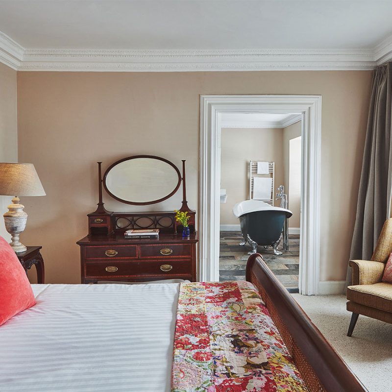 A stunning double bedroom with antique furniture leading into a bathroom with a roll top bath in a Superior Room in Brambles of Inveraray Hotel in Scotland